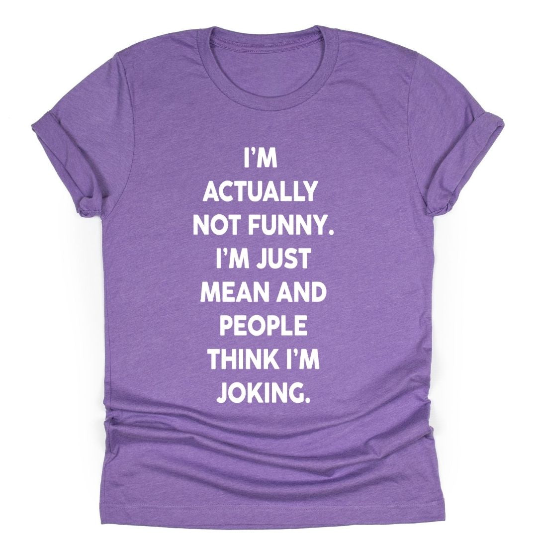 Purple unisex t-shirt with bold white text that reads: ‘I’m actually not funny. I’m just mean and people think I’m joking.’ Shirt laid flat with rolled sleeves.