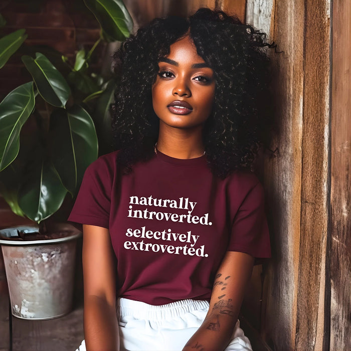 Woman wearing a maroon t-shirt with text, standing next to a plant and wooden wall.